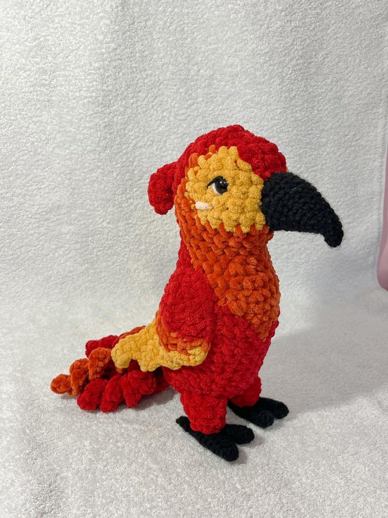 May include: A handmade crochet parrot in vibrant red, orange, and yellow. The plush bird has a black beak and feet, with a detailed tail and head. The soft, textured yarn creates a cuddly appearance, perfect for a child's toy or home decor.