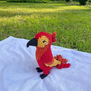 May include: A handmade crochet bird in vibrant red, orange, and yellow hues. The bird has a black beak and feet, and a textured appearance. The bird is sitting on a white surface, with a grassy background.