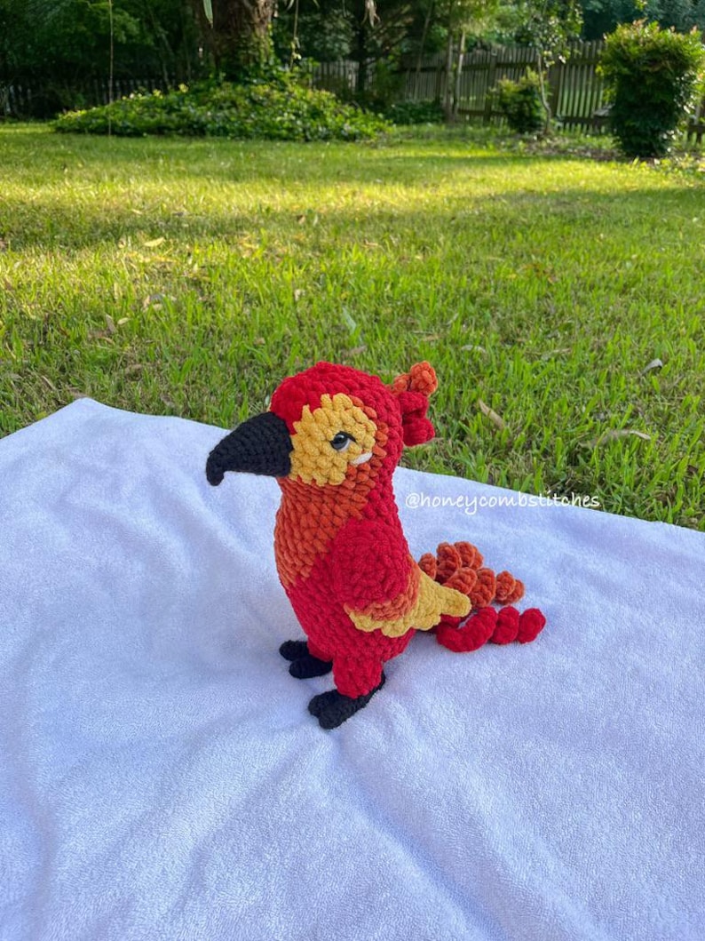 May include: A handmade crochet bird in vibrant red, orange, and yellow hues. The bird has a black beak and feet, and a textured appearance. The bird is sitting on a white surface, with a grassy background.