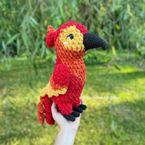 May include: A handmade crochet bird in vibrant red, orange, yellow, and black. The bird has a detailed design with a black beak and feet. The text "honeycombstitches" is visible on the right side. A unique, colorful, and soft plush toy.