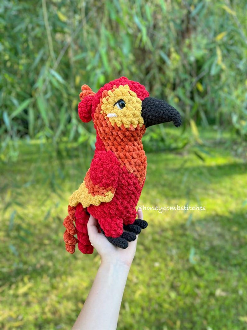 May include: A handmade crochet bird in vibrant red, orange, yellow, and black. The bird has a detailed design with a black beak and feet. The text "honeycombstitches" is visible on the right side. A unique, colorful, and soft plush toy.