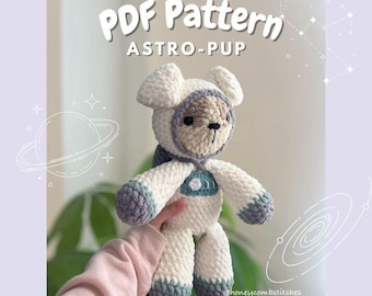 May include: A crocheted plush toy in the shape of a dog wearing an astronaut suit. The toy is white with grey accents and has a brown nose and eyes. The toy is being held by a hand in a pink sweater. The background is a light purple with a white outline of a galaxy.