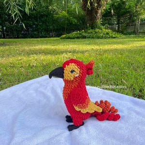 May include: A handmade crochet bird in shades of red, orange, and yellow, with a black beak and feet. The bird is sitting on a white surface, with a green grassy background. The text "honeycombstitches" is visible.