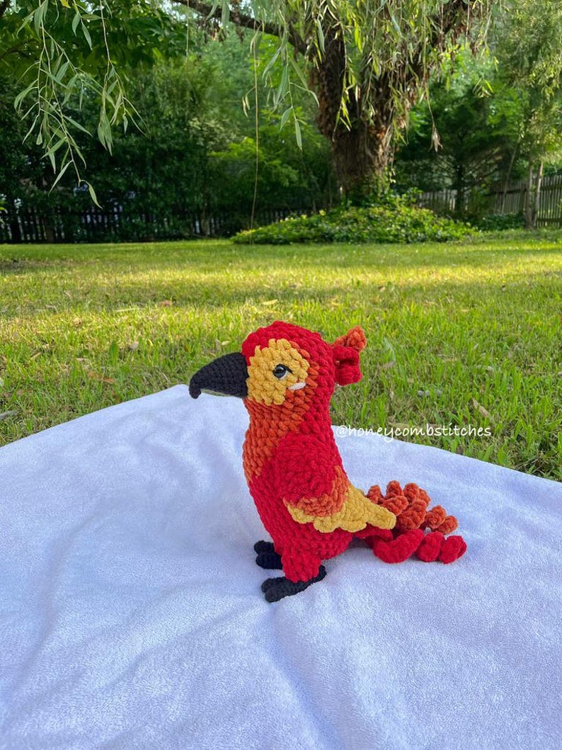 May include: A handmade crochet bird in shades of red, orange, and yellow, with a black beak and feet. The bird is sitting on a white surface, with a green grassy background. The text "honeycombstitches" is visible.