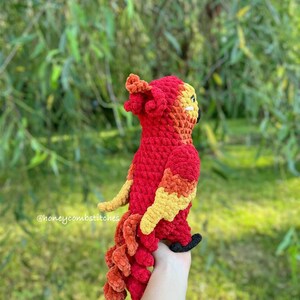 May include: A handmade crochet phoenix in vibrant red, orange, and yellow. The bird has a detailed design with a textured body, wings, and tail. The phoenix is held against a blurred green background. The text "@honeycombstitches" is visible.