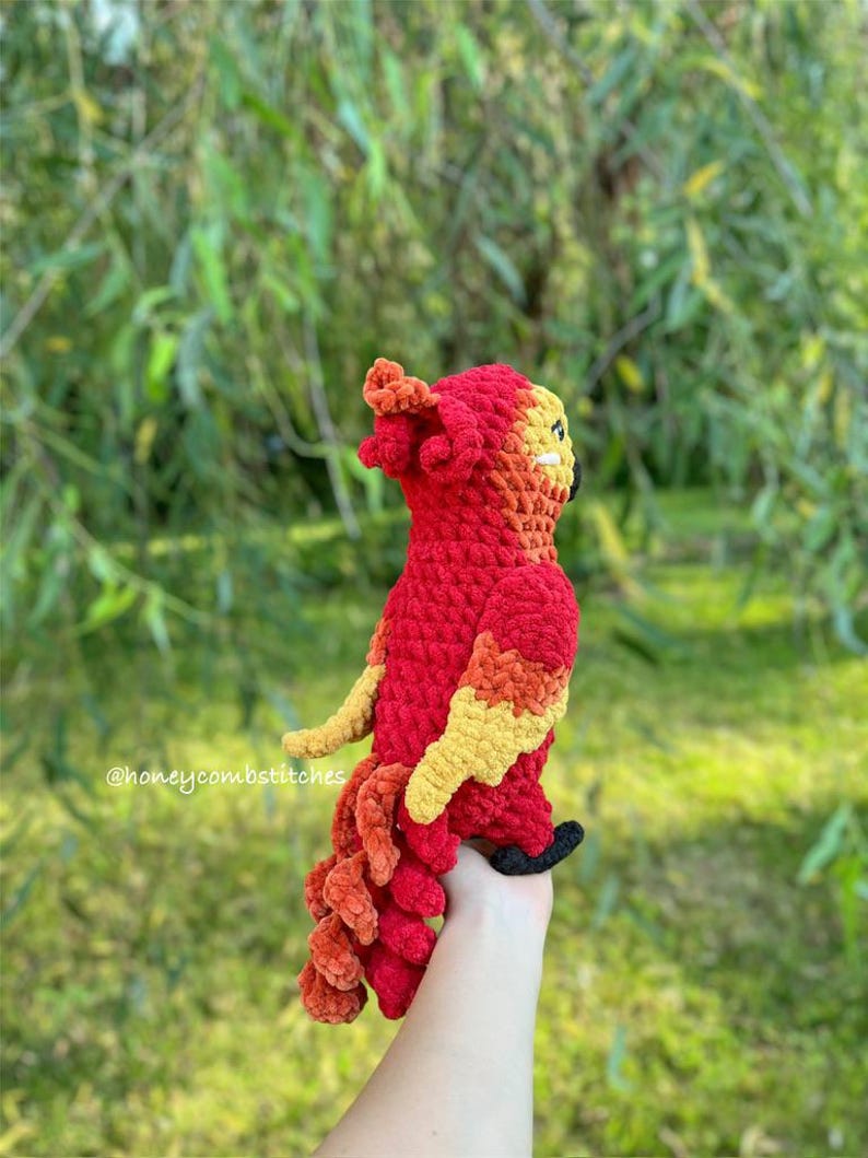 May include: A handmade crochet phoenix in vibrant red, orange, and yellow. The bird has a detailed design with a textured body, wings, and tail. The phoenix is held against a blurred green background. The text "@honeycombstitches" is visible.