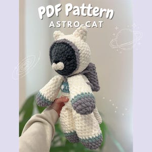 May include: A crocheted Astro-Cat plushie in cream, gray, and blue. The cat is wearing an astronaut helmet and backpack. The text "PDF Pattern ASTRO-CAT" is at the top of the image. The plushie is approximately 10 inches tall.