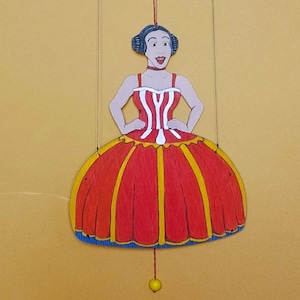 Puede incluir: Una marioneta plana de madera que representa una figura con cabello oscuro recogido en moños, vistiendo un corpiño a rayas rojas y blancas y una falda roja amplia con detalles verticales amarillos. La marioneta cuelga de hilos y tiene una cuenta amarilla.