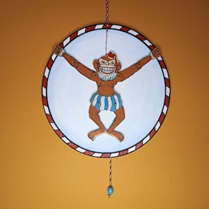 Puede incluir: Un juguete de madera vintage con un mono marrón con una amplia sonrisa, que lleva un sombrero rojo y blanco y pantalones cortos a rayas azules y blancas. El mono está suspendido de una cuerda, sobre un círculo blanco con un borde a rayas rojas y blancas.