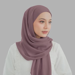 Op de afbeelding: Een mauvekleurige hijab, elegant gedrapeerd, die het hoofd en de schouders bedekt. De stof heeft een zachte, vloeiende uitstraling. De persoon draagt een witte blouse met kanten details.
