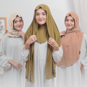 May include: Three women wearing hijabs in different colors. The hijabs are in shades of beige, olive green, and peach. They are all wearing white dresses. The hijabs are draped over their heads and shoulders.
