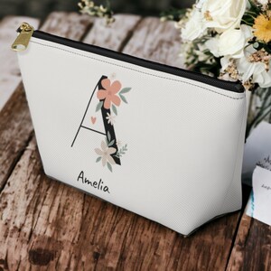 May include: White zippered makeup bag with a black letter A decorated with pink and white flowers. The name Amelia is printed below the letter.