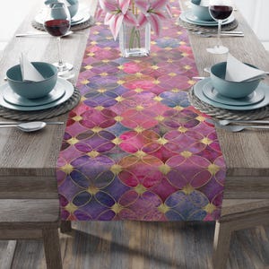 May include: A decorative table runner with a watercolor design. The runner features a pattern of overlapping circles in shades of pink, purple, and blue, accented with gold lines. The runner is placed on a wooden table set for a meal.