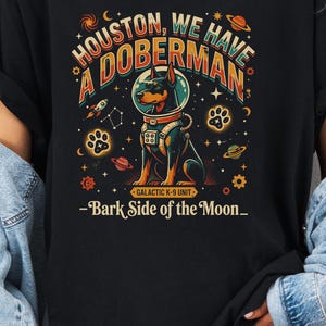 May include: Black t-shirt with a colorful graphic featuring a Doberman Pinscher in an astronaut helmet. Text reads "Houston, We Have A Doberman" and "-Bark Side of the Moon_". The design includes planets, stars, and paw prints.