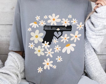 Può includere: Maglietta grigia con una silhouette nera di una pistola con un cuore al centro e il testo "GIRL POWER" sul cannone. La pistola è circondata da margherite bianche con centri gialli.