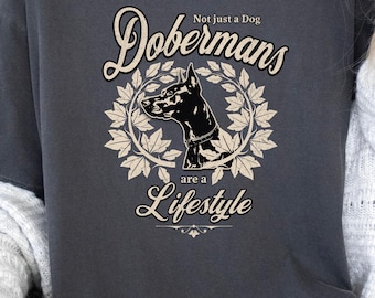 Può includere: T-shirt grigio scuro con un'illustrazione di Dobermann in bianco e nero all'interno di una ghirlanda. La maglietta mostra il testo "Dobermans are a Lifestyle" e "Not just a dog". Un capo d'abbigliamento casual e comodo.
