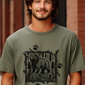 Pu&ograve; includere: T-shirt verde oliva con grafica nera di un orso in una scena forestale, incorniciata da rami e pini. Il design include impronte di zampe di orso. La maglietta &egrave; realizzata in un materiale morbido e confortevole.