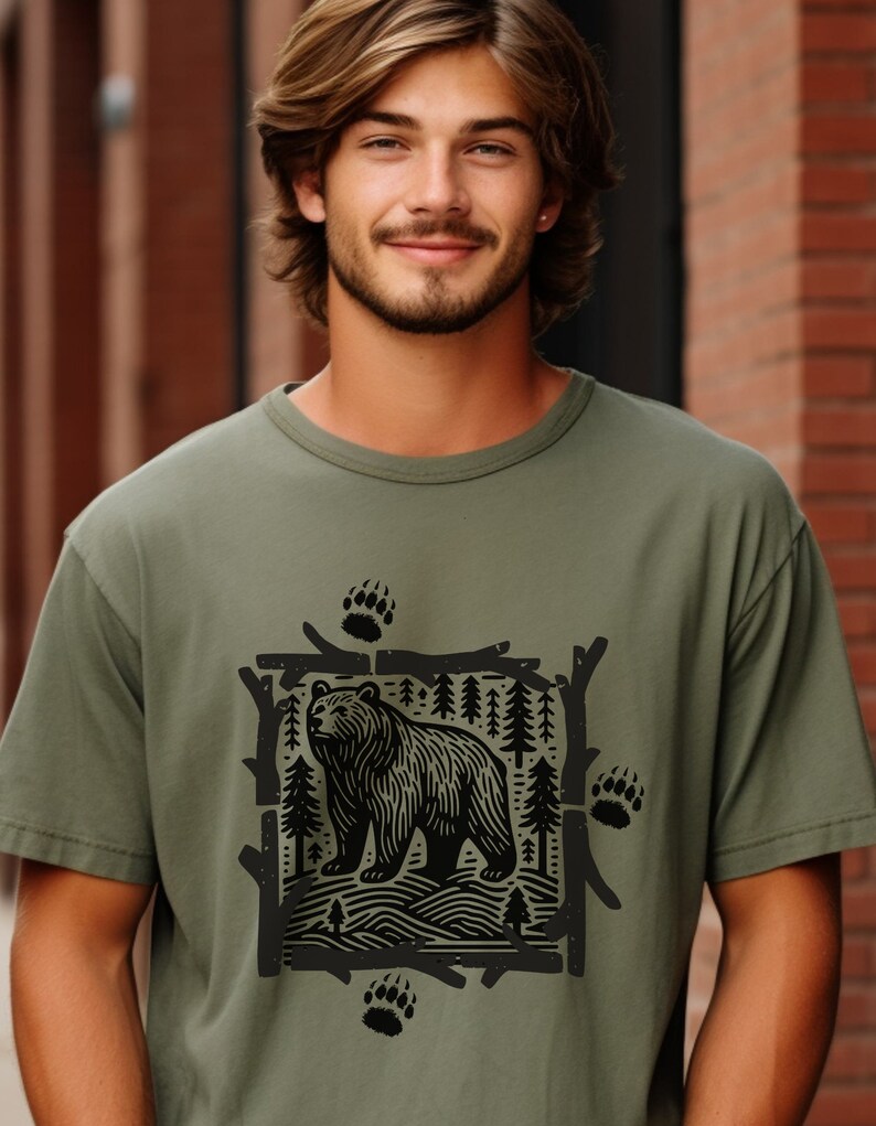 Pu&ograve; includere: T-shirt verde oliva con grafica nera di un orso in una scena forestale, incorniciata da rami e pini. Il design include impronte di zampe di orso. La maglietta &egrave; realizzata in un materiale morbido e confortevole.