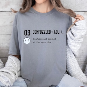 May include: Gray t-shirt with the text "03 CONFUZZLED-(ADJ.)" and a cartoon face with a confused expression. The text below the face reads "Confused and puzzled at the same time."