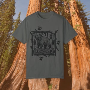 Pu&ograve; includere: T-shirt grigio scuro con una grafica nera di un orso in una scena forestale. Il design &egrave; incorniciato da rami e include impronte di zampe di orso. La maglietta &egrave; a girocollo.