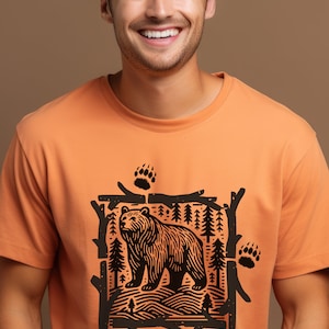 Pu&ograve; includere: T-shirt arancione girocollo con grafica nera di un orso in una cornice di foresta, con impronte di zampe di orso. Il design &egrave; centrato sulla maglietta, creando un'estetica a tema natura.