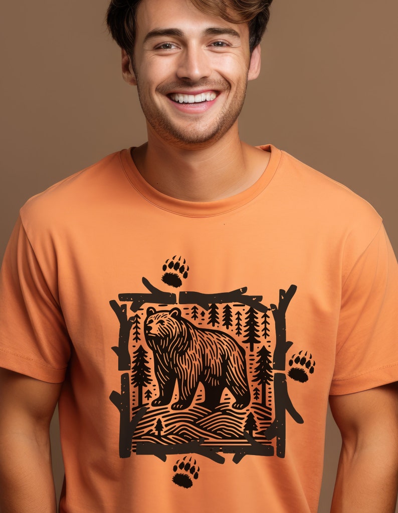Pu&ograve; includere: T-shirt arancione girocollo con grafica nera di un orso in una cornice di foresta, con impronte di zampe di orso. Il design &egrave; centrato sulla maglietta, creando un'estetica a tema natura.