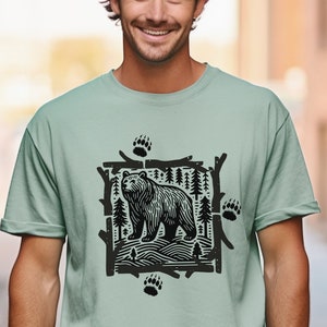 Pu&ograve; includere: T-shirt verde chiaro con una grafica in bianco e nero di un orso in una scena forestale. Il design include impronte di zampe di orso e alberi stilizzati. La maglietta &egrave; realizzata in materiale morbido e ha maniche corte.