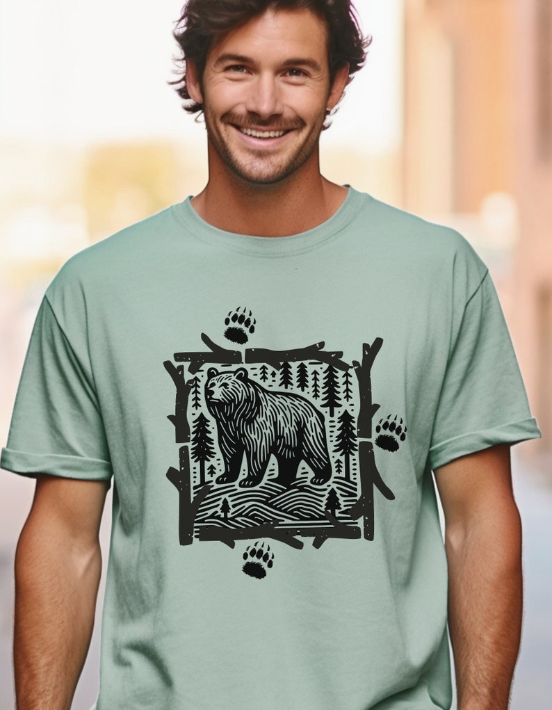 Pu&ograve; includere: T-shirt verde chiaro con una grafica in bianco e nero di un orso in una scena forestale. Il design include impronte di zampe di orso e alberi stilizzati. La maglietta &egrave; realizzata in materiale morbido e ha maniche corte.