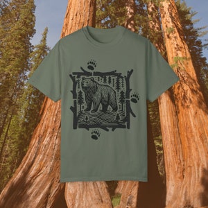 Pu&ograve; includere: T-shirt verde oliva con grafica nera di un orso in una foresta, incorniciata da rami e impronte di zampe. Il disegno &egrave; centrato, con girocollo e maniche corte. Un capo di abbigliamento casual.