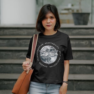 May include: Black t-shirt with a white graphic of a dragonfly on a moon with the words "Change, Transformation, Adaptability, Self-Realization" around the moon.