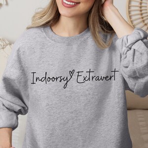 Può includere: Felpa grigia con il testo "Indoorsy Extravert" in lettere nere con un simbolo di cuore tra le parole.