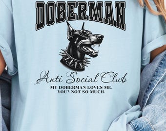 Può includere: T-shirt azzurra con grafica in bianco e nero di un Dobermann Pinscher e la scritta "DOBERMAN" sopra. Sotto c'è "Anti Social Club" e una citazione umoristica. Un capo di abbigliamento casual.