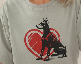 Può includere: Una maglietta grigia con un'illustrazione in bianco e nero di un Dobermann seduto davanti a un cuore rosso. Il cuore ha un aspetto usurato e vintage.