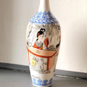 Puede incluir: Un jarrón alto de cerámica blanca con cuello esbelto y borde acampanado. El jarrón presenta patrones florales azules pintados a mano en la parte superior e inferior. El cuerpo del jarrón representa una escena con figuras con vestimenta tradicional, una mesa y un árbol.