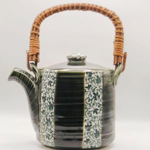 May include: A ceramic teapot with a woven handle. The teapot is dark green with a floral pattern in white and green. The handle is made of woven brown material. The lid has a small knob.