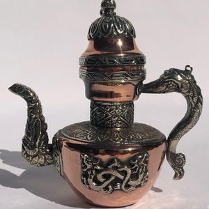 May include: A copper teapot with a silver lid and handle. The teapot is decorated with intricate floral and geometric designs. The handle is shaped like a dragon's head.
