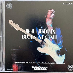 Könnte beinhalten: Eine CD-Hülle mit Jimi Hendrix. Das Albumcover hat einen dunklen Hintergrund mit einem Foto von Hendrix, der eine weiße Gitarre spielt. Der Text "Jimi Hendrix Blues at Cafe" ist deutlich sichtbar. Die CD-Hülle ist aus klarem Kunststoff.