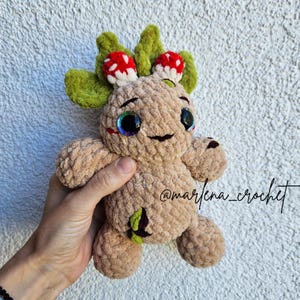 Pu&ograve; includere: Un peluche all'uncinetto color marrone chiaro a forma di creatura arborea con grandi occhi iridescenti, capelli a forma di foglie verdi e due piccoli funghi rossi a pois bianchi sulla testa. Il peluche ha una finitura in filato testurizzato e il testo @marlena_crochet.