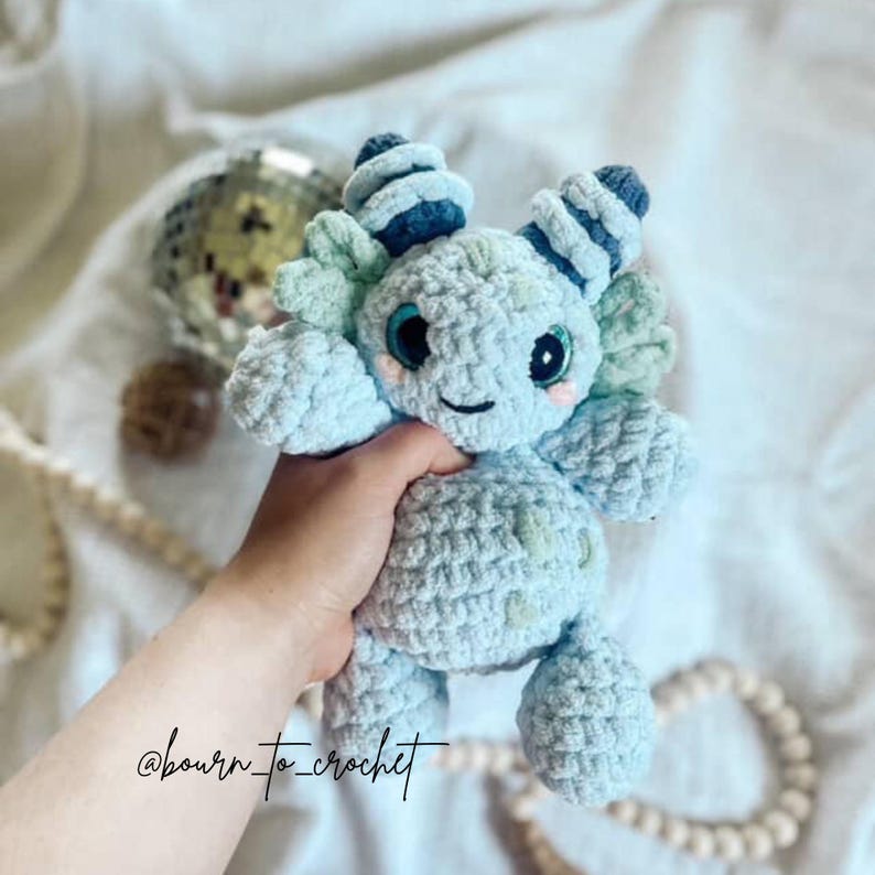 Pu&ograve; includere: Un peluche axolotl all'uncinetto color azzurro con corna a righe blu scuro, grandi occhi neri e guance rosa. Il peluche presenta una struttura in filato morbido e testurizzato con corpo arrotondato, braccia e gambe piccole. Il testo "bourn_to_crochet" &egrave; visibile.