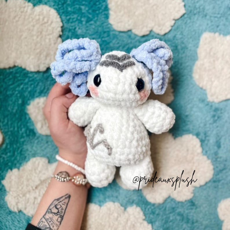 Pu&ograve; includere: Un peluche all'uncinetto bianco con capelli ricci azzurri, occhi scuri e guance rosa. La figura presenta un simbolo grigio ricamato sulla fronte e un disegno grigio ricamato sul petto. Il peluche ha una finitura in filato morbido e testurizzato.