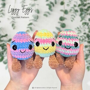 Pode incluir: Três brinquedos de pelúcia de crochê em forma de ovo com pernas marrons, olhos de segurança pretos e sorrisos bordados. O ovo da esquerda é azul e roxo, o central é amarelo e verde, e o da direita é azul-petróleo e rosa. Texto: Leggy Eggs Crochet Pattern.