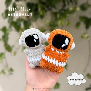 May include: Two small crocheted astronaut figures, one in light gray and one in orange, each featuring a black visor with white highlights and a white band around the midsection. The text Itty Bitty ASTRONAUT and PDF Pattern are visible.