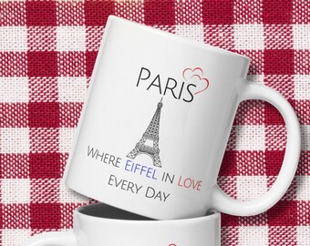 May include: Two white ceramic mugs with a black and white illustration of the Eiffel Tower and the text "Paris where Eiffel in love every day". The mugs are on a red and white checkered tablecloth.
