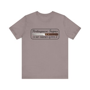 Könnte beinhalten: Ein heidefarbenes T-Shirt mit einem weißen Rechteck und schwarzem Text, der "Portuguese Tapas Zero Facas Given" lautet. Ein Messer mit braunem Griff befindet sich im Rechteck.