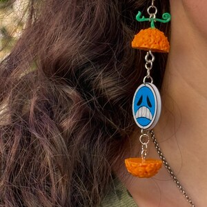 Op de afbeelding: Hangende oorbellen met een groen en oranje ontwerp. De oorbellen hebben een groen bovenstuk, een oranje, getextureerde halve bol, een blauw en wit ovaal met een verdrietig gezicht en nog een oranje halve bol. Ze zijn verbonden door zilveren kettingen.