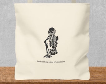 May include: A cream-colored canvas tote bag featuring a black graphic of a skeleton sitting in a hunched position. Below the skeleton, the text reads, The mortifying ordeal of being known. The bag has two matching cream-colored fabric shoulder straps.