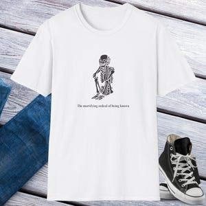 Könnte beinhalten: Weißes T-Shirt mit einem schwarzen Skelett-Design und dem Text "The mortifying ordeal of being known". Ein schwarzer High-Top-Sneaker ist im Bild zu sehen.