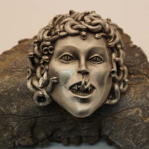 Könnte beinhalten: Eine silberfarbene Skulptur von Medusa, der griechischen mythologischen Figur mit Schlangenhaaren, mit einem bedrohlichen Ausdruck. Die Skulptur ist vor einem rauen, grauen Felsgrund platziert.