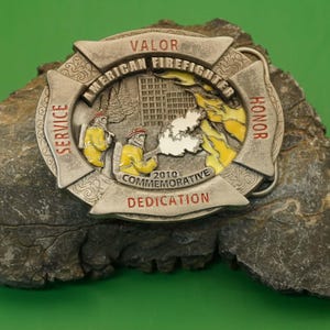 Könnte beinhalten: Eine Gedenkschnalle mit einem Feuerwehr-Malteserkreuz-Design. Die Schnalle trägt die Worte "Valor", "American Firefighter", "Honor", "Service", "2010 Commemorative" und "Dedication". In der Mitte sind Feuerwehrleute und ein brennendes Gebäude abgebildet.