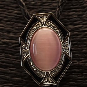 Könnte beinhalten: Eine Bolo-Krawatte mit geometrischem Design. Der silberfarbene Metallanhänger hat einen ovalen, rosabraunen Stein, der von schwarzem Emaille und dekorativen Verzierungen umrahmt ist. Die Krawatte hat eine geflochtene schwarze Kordel.